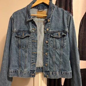 Klassisk blå jeansjacka med knappar - Snygg blå jeansjacka från 157 med klassisk passform och silverfärgade knappar framtill. Jackan har två bröstfickor med lock och knapp, samt långa ärmar. Perfekt att slänga över en hoodie eller t-shirt för en chill look.