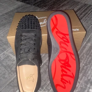 Christian Louboutin svarta sneakers  - Svarta sneakers från Christian Louboutin med coola svarta nitar på tån och klassisk röd sula med logga. Skorna har snörning och är gjorda i mocka med rund tå. Medföljer dustbag och extra nitar. Perfekt för dig som vill sticka ut med lyxig streetstyle.