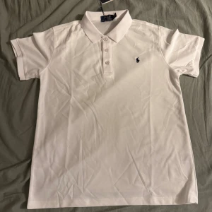 Vit pikétröja från Polo Ralph Lauren - Klassisk vit pikétröja från Polo Ralph Lauren med korta ärmar, krage och knappar framtill. Tröjan har den ikoniska broderade loggan i svart på bröstet och är tillverkad i bomull för en skön och stilren look.