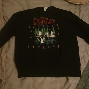 Svart jultröja Christmas Things - Svart tröja med tryck inspirerat av Stranger Things och texten 'Christmas Things'. Motiv med cyklande figurer, julgranar, snöflingor och färgglada ljusslingor. Perfekt för dig som gillar nördiga jultröjor och popkultur.