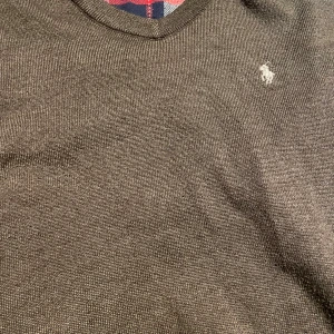 Brun stickad tröja Polo Ralph Lauren - Snygg brun stickad tröja från Polo Ralph Lauren med v-ringad hals och klassisk logga broderad på bröstet. Tröjan har quiltade mocka armbågslappar som ger en schysst touch. Perfekt för dig som gillar stilrena detaljer och tidlös design.