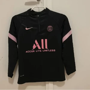 PSG träningströja från Nike - Snygg svart Paris Saint-Germain fotbollströja från Nike med rosa detaljer och logga. Tröjan har lång ärm, halv dragkedja och rosa tryck på bröstet. perfekt för träning. Tröjan är i storlek 158cm.