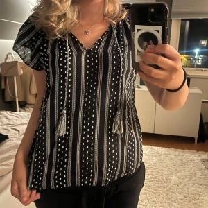Mönstrad blus - Säljer denna sååå fina och söta svara blus med vita ränder och prickar, v-ringad med dekorativa snören och tofsar framtill. Verkligen såå fin🦨🪐