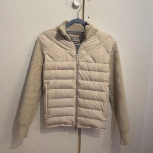 Beige quiltad pufferjacka Moncler - Snygg beige pufferjacka från Moncler med stickade ärmar och hög krage. Jackan har quiltad framsida, dragkedja med Moncler-logga och ett märke på ärmen. Perfekt för dig som gillar stilren design och exklusiva detaljer.