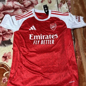 Arsenal Gyökeres 14 Adidas fotbollströja - Säljer en officiell Arsenal hemmatröja från Adidas med Gyökeres och nummer 14 på ryggen. Tröjan är röd med vita detaljer och har ett subtilt mönster över hela framsidan. Kortärmad modell i lätt och ventilerande material, perfekt för match eller träning.