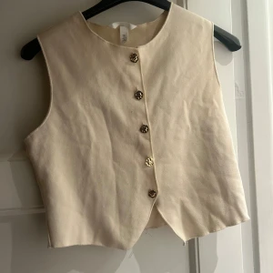 Beige väst med guldfärgade knappar - Snygg beige väst från H&M i storlek S. Västen har en rak och stilren form med fem guldfärgade knappar framtill. Materialet känns mjukt och har en lätt struktur. Perfekt att styla över en t-shirt eller skjorta för en cool look.