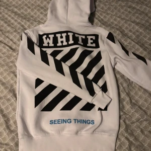 Vit Off-White hoodie XS med tryck - Säljer en vit hoodie från Off-White i storlek XS med ikoniska svarta ränder och texten 'WHITE' på ryggen samt 'SEEING THINGS' i blått. Tröjan har huva med snörning, känguruficka och coolt tryck på ärmarna. Perfekt för dig som gillar streetwear och vill sticka ut.