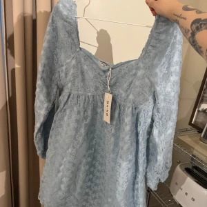 Blommig ljusblå klänning från NA-KD - Supersöt ljusblå klänning från NA-KD med blommigt spetsmönster och långärmade puffärmar. Klänningen har en fyrkantig ringning och är kort i modellen - oanvänd. 