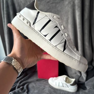 Valentino Garavani vita sneakers - Snygga vita sneakers från Valentino Garavani med stor svart VLTN-logga på sidan och coola nitar vid hälen. Skorna har rund tå, platt sula och snörning. Hälpartiet är metallic-silver och materialet är lyxigt skinn. Perfekt för dig som gillar statement pieces.