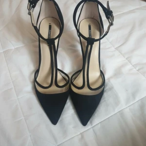 Svarta pumps med spetsig tå från Zara - Svarta pumps från Zara Basic med smala remmar och snyggt spänne runt ankeln. Skorna har spetsig tå och hög klack, vilket ger en elegant siluett. Materialet är mocka och insidan är ljusbeige. Perfekt för dig som gillar stilrena och trendiga skor. Oanvända 