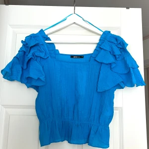 Pastellblå volangblus Gina  - Snygg blå topp från Gina Tricot med volangdetaljer på ärmarna. Toppen har en fyrkantig halsringning och korta ärmar. Perfekt för en somrig look! Nypris 359