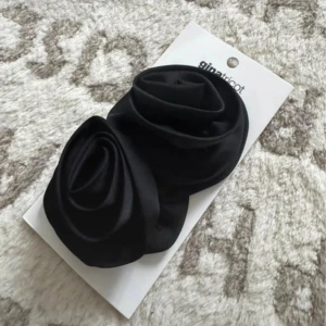 Svart diadem med rosor från Gina Tricot - Snyggt svart diadem från Gina Tricot med två stora tygrosor som ger en cool statement-look. Perfekt accessoar för att lyfta din outfit och ge en trendig vibe. Diademet är gjort i ett slätt tyg som känns mjukt mot håret.