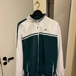 Vit och grön vindjacka från Lacoste - Snygg vindjacka från Lacoste i vitt och mörkgrönt med dragkedja framtill, ståkrage och broderad krokodil-logga på bröstet. Jackan har två fickor med dragkedja och elastisk nederkant. Perfekt för dig som gillar sportig och clean stil. Jag säljer även byxorna till denna jacka. Se mina andra annonser. 