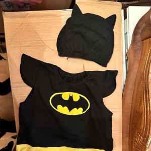 Batman klänning med öronmössa - Supersöt svart body för små barn med Batmans logga/motiv i gult framtill och matchande svart mössa med öron. Bodyn har korta ärmar, resår i halsen och en gul rand nertill. Perfekt för alla små superhjältar! Från cirka 6mån uppåt. Svart Batman body med en gul Batman-logga på framsidan och en matchande mössa med öron. Perfekt för maskerad eller varför inte halloween 👻 party för de små eller födelsedag 🎉 ? 