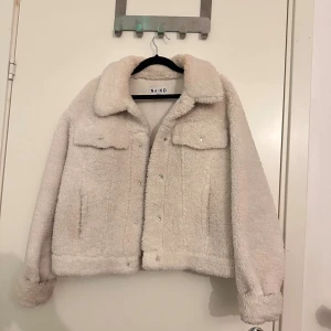 Vit teddyjacka från NA-KD - Mysig vit teddyjacka från NA-KD med krage och tryckknappar framtill. Jackan har två bröstfickor med lock och långa ärmar. Perfekt för att hålla sig varm och se stylish ut under kyliga dagar. Fleece-liknande material ger en fluffig och skön känsla.