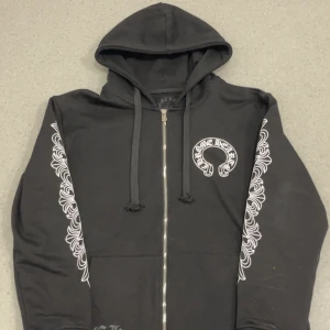 Svart Chrome Hearts hoodie med tryck - Svart hoodie från Chrome Hearts med vita tryck längs ärmarna och stort logotryck på ryggen. Tröjan har huva, snörning och dragkedja framtill. Grym streetwear vibe och riktigt snygga detaljer.