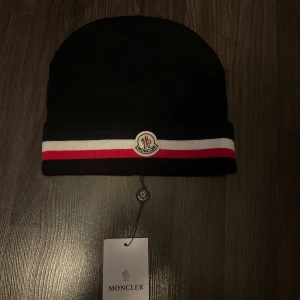 Svart Moncler mössa med ränder - Svart mössa från Moncler med bred uppvikt kant och klassisk logga framtill. Kant med marinblå, vit och röd rand ger en snygg kontrast. Stickad i mjukt material som passar perfekt för kyliga dagar. En stilren accessoar för dig som gillar exklusiva märken.