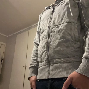 Grå hoodie från BOSS  - Säljer en grå hoodie från BOSS med vit piping längs ärmar och bröst. Tröjan har huva med snörning, dragkedja framtill och två fickor. Perfekt för chill dagar eller när du vill ha en clean streetstyle-look.