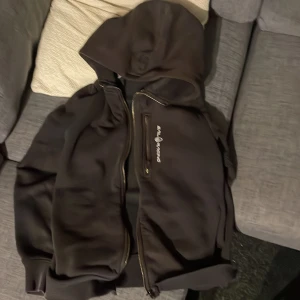 Svart zip tröja från Sail Racing - Svart zip tröja från Sail Racing med huva och dragkedja framtill. Jackan har en bröstficka med dragkedja och vit logotyp på framsidan. Materialet är mjukt och känns som syntet, perfekt för blåsiga dagar. Passformen är normal och tröjan har långa ärmar.