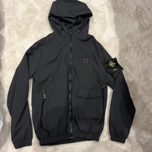 Svart vindjacka från Stone Island - Svart vindjacka från Stone Island med huva, dragkedja framtill och elastiska muddar. Jackan har en ikonisk patch på ärmen, två fickor med dragkedja och en ficka med lock. Materialet är lätt och syntetiskt, perfekt för blåsiga dagar.