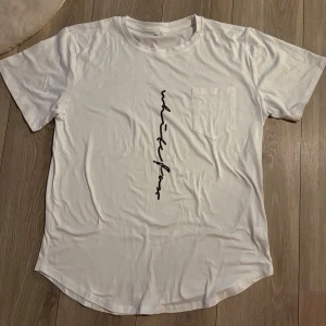 Vit t-shirt med svart tryck XL - Snygg vit t-shirt i storlek XL med svart vertikalt tryck på framsidan och en bröstficka. Rundad nederkant och klassisk passform. Mjuk och skön känsla, perfekt till jeans eller shorts. Materialet känns som bomull.