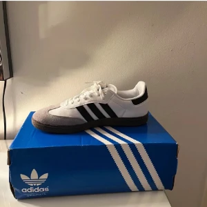 Adidas Samba sneakers vit/svart - Adidas Samba sneakers med klassisk design i vitt och svart. Skorna har svarta ränder på sidorna, grå mockadetaljer vid tån och snörning. Yttermaterialet är en mix av skinn och mocka, med platt sula och rund tå. Perfekt för dig som gillar retrostil och streetwear.