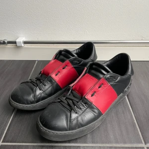 Valentino Garavani svarta och röda sneakers - Snygga svarta och röda sneakers från Valentino Garavani med coola nitar på hälen. Skorna har snörning, rund tå och är tillverkade i skinn. Den röda panelen ger en fet kontrast mot det svarta och gör dem riktigt unika. Perfekta för dig som vill sticka ut med designer på fötterna. Givetvis äkta🎯
