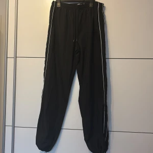 Svarta täckbyxor från FB Siser M - Svarta täckbyxor från FB Siser i storlek M med vita ränder längs sidorna. Byxorna har elastisk midja med snörning och resår vid bensluten för en skön passform. Perfekta för dig som gillar sportig stil och vill ha bekväma byxor med snygga detaljer.