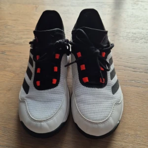 Adidas Adizero Ubersonic tennisskor - Säljer ett par Adidas Adizero Ubersonic tennisskor i vitt och svart med röda detaljer. Skorna har mesh-ovandel för bra ventilation, snörning och slitstark sula med bra grepp. Perfekta för dig som vill ha snabba och lätta skor på tennisbanan.
