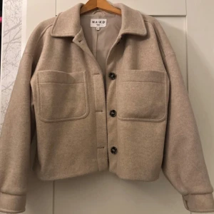 Beige kappa från NA-KD - Snygg beige kappa från NA-KD med stora bröstfickor och knappar framtill. Jackan har en loose passform, klassisk krage och långa ärmar med knapp vid mudden. Perfekt att slänga på över en hoodie eller t-shirt för en chill look.