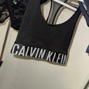 Svart linnetopp från Calvin Klein - Svart linnetopp från Calvin Klein med bred resår och vit logotyp längs nederkanten. Toppen har en sportig, croppad passform och ärmlös design med rundad halsringning. Perfekt för dig som gillar stilrena och bekväma plagg med tydlig branding.