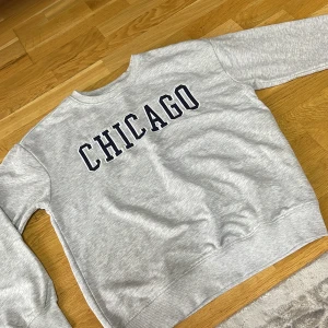 Grå sweatshirt med CHICAGO-tryck - Snygg grå sweatshirt med stort CHICAGO-tryck i svart på bröstet. Tröjan har rund hals, ribbade muddar vid ärmslut och nederkant samt långärmad passform. Helt ny