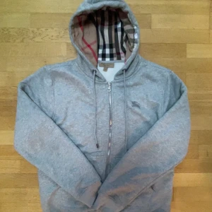 Grå Burberry hoodie med dragkedja - Passar mer som M!  Snygg grå hoodie från Burberry med klassiskt rutigt foder i huvan och broderad logga på bröstet. Tröjan har dragkedja framtill, snörning vid huvan och långa ärmar. Perfekt för dig som gillar stilrena och exklusiva streetwear-plagg. Prislapp är kvar