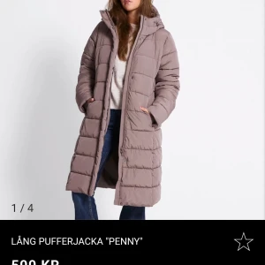 Lång beige pufferjacka Penny HELT NY!!! - Lång, figursydd pufferjacka i beige med huva och bred mudd vid ärmslut. Jackan har tvåvägsdragkedja, tryckknappar och fickor med dragkedja. Huva med dragsko och vattenavvisande yta. Perfekt för kalla dagar och snygg till jeans. HELT NY!! ALDRIG ANVÄND! 