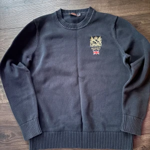 Mörkblå stickad tröja från Morris - Snygg mörkblå stickad tröja från Morris med broderad guldfärgad logga och brittisk flagga på bröstet. Tröjan har rund hals, ribbade muddar och är långärmad. Perfekt för dig som gillar klassisk och stilren look. Lappen har lossnat lite men inget man märker, Hör av dig vid frågor!!
