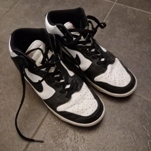 Nike Dunk High svart/vit sneakers - Nike Dunk High sneakers i svart och vitt med klassisk Swoosh på sidan. Skorna har högt skaft, snörning och rund tå. Ovandelen är i skinn och syntet, med perforeringar på tån för extra ventilation. Perfekt för dig som gillar retrostil och streetwear. 