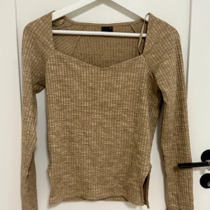 Beige ribbad långärmad topp - Säljer en beige långärmad topp med ribbad struktur och rutigt mönster. Toppen har en snygg v-ringad hals och slits på sidan för extra detalj. Materialet känns mjukt och stretchigt, perfekt för dig som gillar en chill och stilren look. Stl M