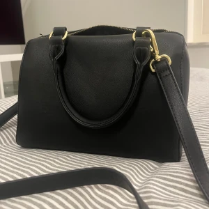 Svart handväska med gulddetaljer - Säljer denna snygga väska som knappt är använd. Den är i mycket bra skick och är mycket rymlig. Den har två inre fack och en axelrem som man kan ta av eller justera som man själv vill💗