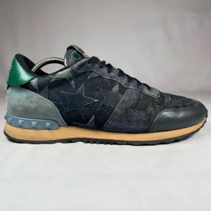 Valentino Garavani Rockrunner sneakers - Snygga Valentino Garavani Rockrunner sneakers i mörkblå mocka och skinn med stjärnmönster och coola nitar på hälen. Skorna har svarta snören, grön detalj bak och beige sula. Perfekta för dig som vill sticka ut med exklusiva och trendiga sneakers. Storlek 41 men eftersom det är valentinos så är det storlek 42!