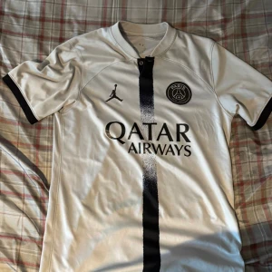 PSG vit fotbollströja Jordan x Qatar - Säljer en snygg PSG fotbollströja i vitt med svart vertikal rand och svarta detaljer på ärmslut. Tröjan har Jordan-loggan och Paris Saint-Germain-emblemet på bröstet samt Qatar Airways tryck. Tillverkad i lätt polyester, perfekt för match eller träning.