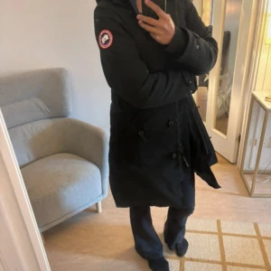 Svart lång dunjacka från Canada Goose - Säljer en svart lång dunjacka från Canada Goose med klassisk patch på ärmen. Jackan har dragkedja, knappar och justerbar huva. Insidan är quiltad och jackan är perfekt för kalla vinterdagar. Snygg och tidlös modell som håller dig varm hela säsongen. Stor i passformen så passar M/L