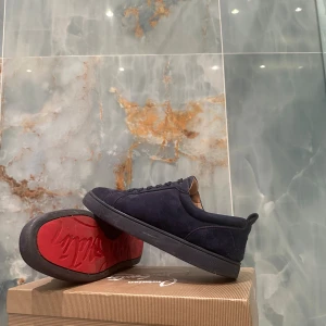 Mörkblå sneakers från Louboutin - Snygga mörkblå sneakers från Christian Louboutin i mocka med klassisk rund tå och platt sula. Insidan är beige och yttersulan är röd med Louboutin-logga. Skorna har snörning och en stilren, minimalistisk design som passar till många outfits.