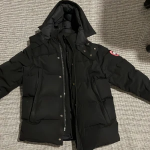 Svart dunjacka från Canada Goose - Säljer en svart dunjacka från Canada Goose med huva och klassisk patch på ärmen. Jackan har dragkedja och knappar framtill, samt stora fickor. Perfekt för kalla vinterdagar och har en riktigt schysst puffer-look.