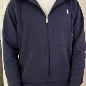Marinblå hoodie från Polo Ralph Lauren - Säljer en marinblå hoodie från Polo Ralph Lauren med dragkedja och klassisk logga broderad på bröstet. Tröjan har huva med snörning, två fickor fram och är tillverkad i mjuk bomullsblandning. Perfekt för en chill och stilren look. 