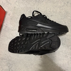 Nike Air Max 90 helsvarta sneakers - Säljer ett par helsvarta Nike Air Max 90 sneakers med klassisk chunky sula och synlig Air Max-dämpning. Skorna har en mix av skinn och syntetmaterial, med snörning och diskret Nike-logga på sidan. Perfekta för dig som gillar en clean och sportig look.