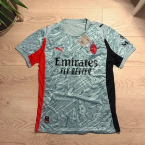 AC Milan kortärmad fotbollströja Puma - AC Milan matchtröja från Puma i ljusblått med abstrakt mönster, svarta och röda paneler på sidorna. Tryckt klubbmärke och Puma-logga på bröstet, samt Emirates Fly Better sponsortryck. Tillverkad i lätt och ventilerande polyester.