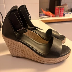 Svarta kilklackade sandaler från XIT - Snygga svarta sandaler från XIT med bred rem över tårna och justerbart spänne vid ankeln. Skorna har hög kilklack i flätad jute som ger en somrig vibe. Ovandelen är i svart syntetmaterial och sulan är platt framtill med öppen tå.