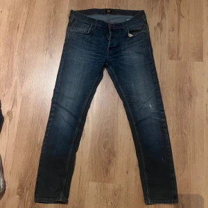 Mörkblå Lee jeans slim fit 34/32 - Snygga mörkblå jeans från Lee med skinny passform och klassisk femficksdesign. Jeansen har subtila slitningar och Lee-logga på bakfickan. Materialet är jeans med stretch för extra komfort. Perfekt för dig som gillar en smal siluett och stilren look. (67)