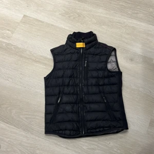 Svart dunväst från Parajumpers - Snygg svart dunväst från Parajumpers med quiltad design och hög krage. Västen har dragkedja framtill, två fickor med dragkedja och en gul detalj vid halsen. Insidan är grå och lätt vadderad för extra värme.