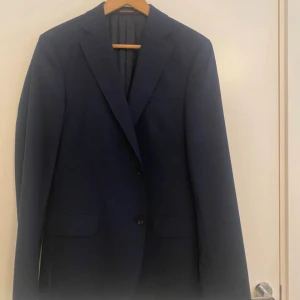 Mörkblå kavaj Oscar Jacobson 48 - Snygg mörkblå kavaj från Oscar Jacobson, modell Edmund Blazer. Tillverkad i Italien med tyg från Vitale Barberis Canonico. Klassisk passform med två knappar framtill och fickor med lock. Perfekt för dig som vill ha en stilren och tidlös look.